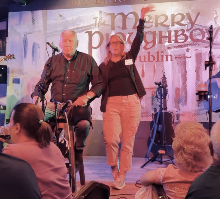 Lisa Solomon with her arm raised on stage at the Merry Ploughboys pub in Dublin, standing beside a seated musician