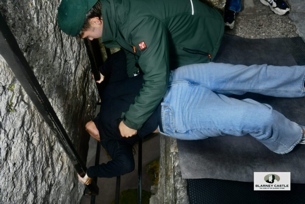 Lisa Solomon leaning back to kiss the Blarney Stone at Blarney Castle in Ireland, held steady by a staff member